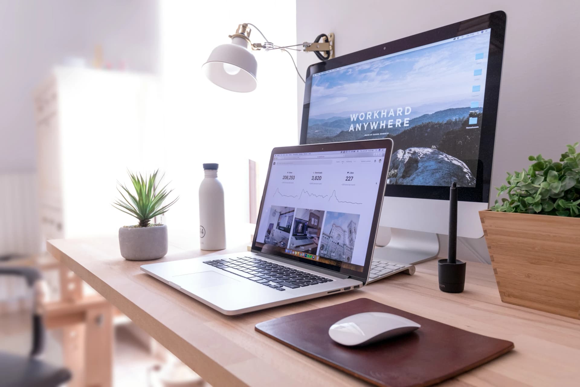 Tidy wooden desk with a laptop showing an analytics dashboard, a desktop monitor reading WORKHARD ANYWHERE, a mouse, and potted plants