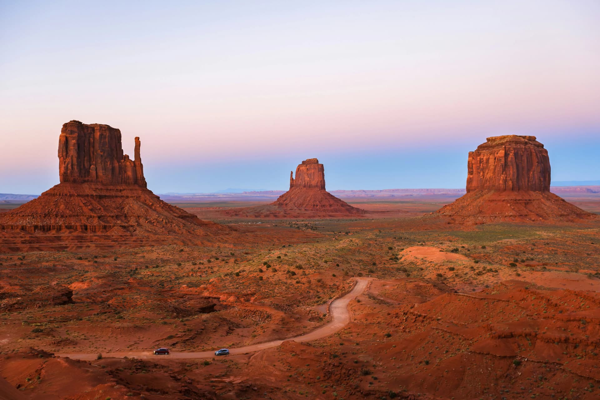 Red sandstone buttes of Monument Valley at sunset, with a dirt road winding through the desert and two cars on it