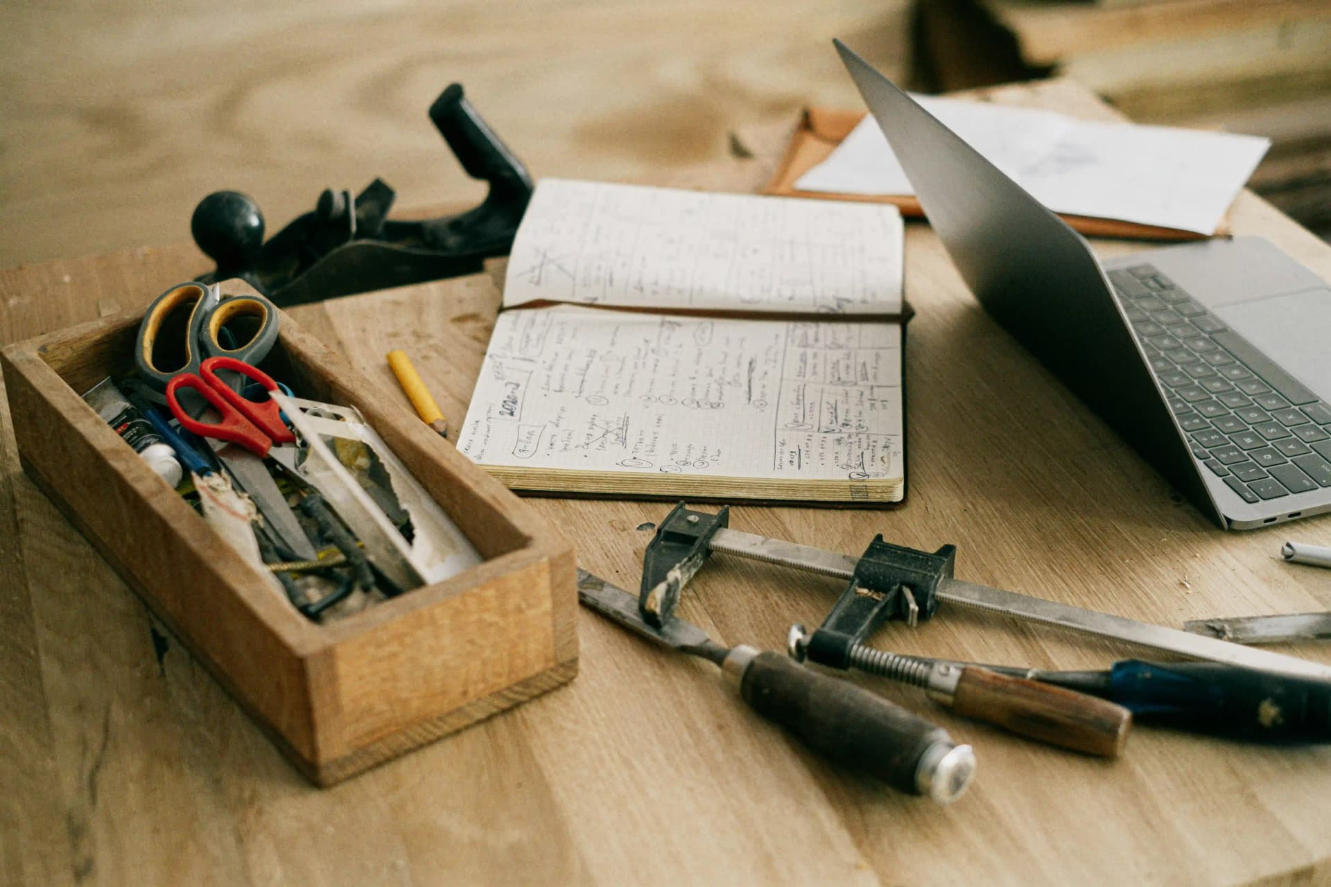 Wooden workbench with a box of scissors and tools, chisels, a bar clamp, a hand plane, an open handwritten notebook, and a laptop